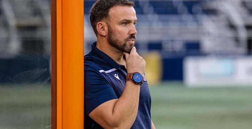 A football coach stands in the technical area looking onto the pitch